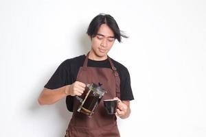 ritratto di attraente asiatico barista uomo nel Marrone grembiule scrosciante caffè in un' tazza a partire dal francese stampa caffè creatore. isolato Immagine su bianca sfondo foto