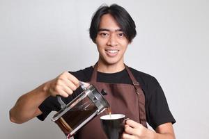 ritratto di attraente asiatico barista uomo nel Marrone grembiule scrosciante caffè in un' tazza a partire dal francese stampa caffè creatore. isolato Immagine su bianca sfondo foto