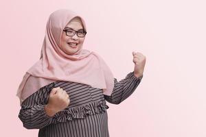 ritratto di allegro asiatico donna con hijab, raccolta il suo cazzotto, festeggiare successo. pubblicità concetto. isolato Immagine su rosa sfondo foto