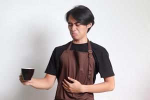 ritratto di attraente asiatico barista uomo nel Marrone grembiule sofferenza a partire dal stomaco dolore potabile un' tazza di caffè. isolato Immagine su bianca sfondo foto