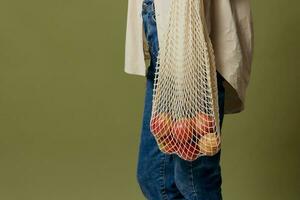 ritagliata tiro di bellissimo sottile femmina nel biancheria casuale camicia hold corda Borsa con frutta su gomito in posa isolato su al di sopra di oliva verde pastello sfondo. eco-friendly naturale prodotti concetto. lato Visualizza foto