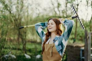 un' donna sorridente meravigliosamente e guardare a il telecamera, un' contadino nel opera Abiti e un grembiule Lavorando all'aperto nel natura e Tenere un' rastrello per raccogliere erba e foraggio per il animali nel il giardino foto