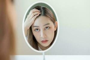 danneggiato capelli, viso grave asiatico giovane donna preoccupazione guardare a cuoio capelluto nel specchio, mano nel rompere in davanti capelli perdita, magro problema sintomo a casa. Salute cura shampoo bellezza, isolato su sfondo. foto