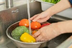 vicino su asiatico giovane donna lavaggio pomodoro, broccoli, carota fresco la verdura, paprica con spruzzo acqua nel bacino di acqua su Lavello nel cucina, preparazione fresco insalata, cucinando pasto. salutare cibo le persone. foto