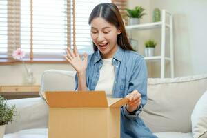 contento emozionato, asiatico giovane donna, ragazza cliente seduta su divano a casa, apertura e disimballaggio cartone scatola scatola di cartone pacco dopo acquisto ordinazione regalo, shopping in linea, consegna servizio concetto. foto