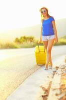 giovane donna con un' giallo valigia è in viaggio su il strada autostop foto