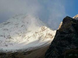 atmosferico paesaggio con acuto rocce e alto nevoso montagna superiore nel bufera di neve Basso nuvole a coperto. drammatico cupo scenario con grande neve montagne e ghiacciaio nel grigio nuvoloso cielo a nevoso tempo metereologico foto