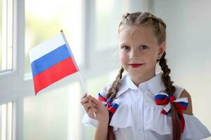 scuola bambino sta e detiene il bandiera di Russia. poco ragazza con viso pittura di russo simbolismo. russo bandiera giorno. foto