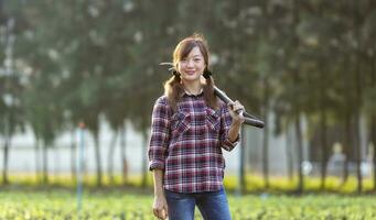 ritratto di asiatico donna contadino è trasporto giardino forchetta mentre in piedi nel sua campagna verdure azienda agricola durante primavera stagione per salutare dieta cibo concetto. foto