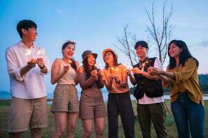 gruppo di giovane asiatico persone siamo godere campeggio , giocando sparkler nel naturale campeggio a crepuscolo foto