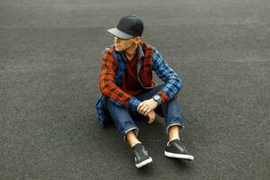 elegante giovane uomo nel un' nero berretto, camicia, jeans e scarpe da ginnastica si siede su il marciapiede. foto