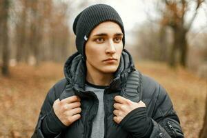 elegante giovane uomo nel un' nero a maglia cappello e giacca in piedi vicino il autunno alberi. foto