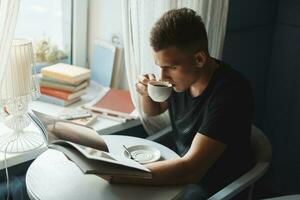 bello giovane tipo con caffè lettura un' libro a il tavolo foto