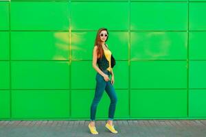 elegante bellissimo donna nel un' giallo maglietta, jeans e scarpe da ginnastica in posa vicino il verde parete. foto