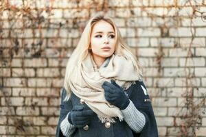 giovane bellissimo bionda donna con alla moda caldo sciarpa vicino un' mattone parete foto