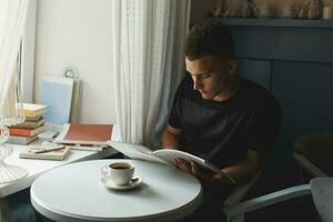 bello giovane uomo lettura un' libro. caffè su il tavolo. foto