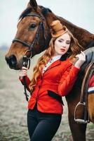 ragazza fantino dai capelli rossi in un cardigan rosso e stivali alti neri con un cavallo