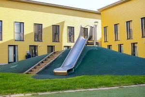 un' metallo diapositiva su un' figli di terreno di gioco con un' verde prato. le scale per il collina. giallo Casa foto
