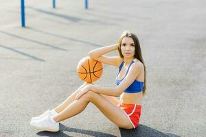 un' giovane atletico donna si siede su il pavimento e detiene un' pallacanestro nel un' gli sport stadio, all'aperto, riposo dopo un' allenarsi foto