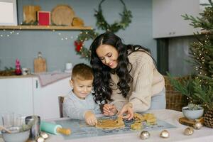 un' contento bellissimo madre con un' giovane figlio è taglio su Natale biscotto muffe a partire dal il Impasto nel il cucina, ridendo. foto