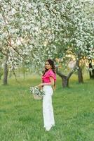 un' bellissimo giovane europeo donna con trecce nel il primavera nel il parco detiene un' cestino di fiori nel un' fioritura frutteto di Mela alberi foto