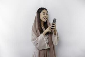 un' contento asiatico musulmano donna indossare un' foulard, Tenere sua Telefono, isolato di bianca sfondo foto