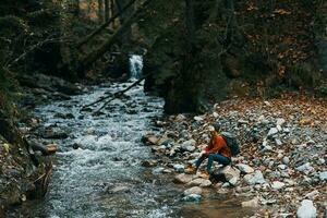 donna viaggiatore vicino un' montagna fiume nel il foresta si siede su il riva autunno paesaggio foto