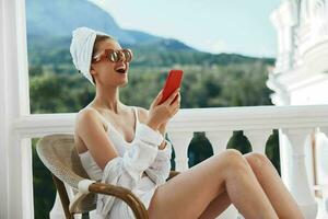 bellissimo donna nel un' bianca camicia si siede su il balcone con un' Telefono rilassamento concetto foto