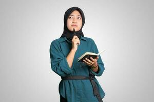 ritratto di eccitato asiatico musulmano donna con hijab scrittura su Nota libro e pensiero con penna su mento. pubblicità concetto. isolato Immagine su bianca sfondo foto