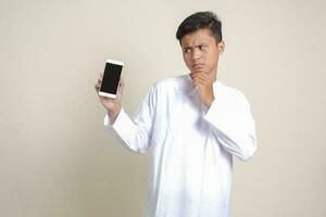 ritratto di attraente asiatico musulmano uomo nel bianca camicia mostrando e presentazione vuoto schermo mobile Telefono. pubblicità concetto. isolato Immagine su grigio foto