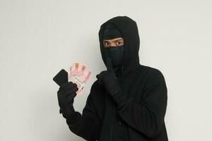 ritratto di misterioso uomo indossare nero felpa con cappuccio e maschera furto uno centinaio mille rupia con successo a partire dal vittima. isolato Immagine su grigio sfondo foto