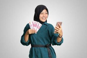 ritratto di eccitato asiatico hijab donna nel casuale attrezzatura mostrando uno centinaio mille rupia mentre Tenere un' mobile Telefono. finanziario e risparmi concetto. isolato Immagine su bianca sfondo foto