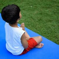 asiatico inteligente ragazzo fare yoga posa nel il società parco all'aperto, figli di yoga posa. il poco ragazzo fare yoga e meditazione esercizio. foto