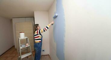 donna vestito nel tuta da lavoro e a strisce camicetta, profilo Visualizza, con un' rullo nel sua mano pittura un' bianca parete con blu dipingere nel un vuoto camera foto