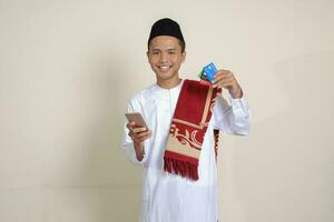 ritratto di attraente asiatico musulmano uomo nel bianca camicia con zucchetto Tenere un' mobile Telefono e presentazione credito carta. isolato Immagine su grigio sfondo foto