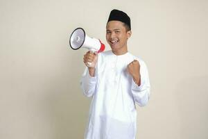 ritratto di attraente asiatico musulmano uomo nel bianca camicia con zucchetto A proposito di più forte utilizzando megafono, promozione Prodotto. pubblicità concetto. isolato Immagine su grigio sfondo foto