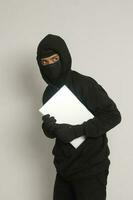 misterioso rapinatore ladro uomo indossare nero felpa con cappuccio e maschera furto il computer portatile e spione fuori. isolato Immagine su grigio sfondo foto