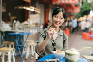 contento giovane asiatico donna zaino viaggiatore godendo strada cibo a Cina cittadina strada cibo mercato nel bangkok, Tailandia. viaggiatore controllo su lato strade. foto