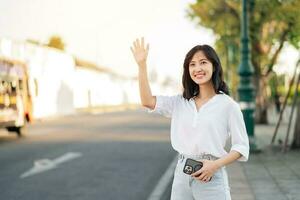 ritratto giovane bellissimo asiatico donna agitando mano per amico di il strada nel soleggiato vacanza. foto