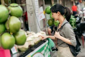 contento giovane asiatico donna zaino viaggiatore In piedi nel davanti di Noce di cocco succo negozio a Cina cittadina strada cibo mercato nel bangkok, Tailandia. viaggiatore controllo su lato strade. foto