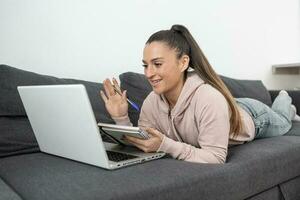 donna agitando a il telecamera su sua il computer portatile mentre assunzione Appunti nel un' taccuino foto