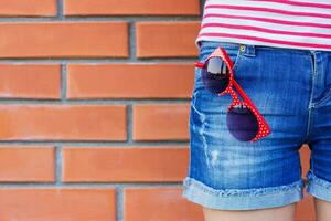 ragazza indossare blu jeans con paio di rosso occhiali da sole nel tasca. moda concetto. foto