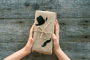 contento padri giorno concetto. bambini è mani hold un' regalo scatola per papà fatto di mestiere carta su un' di legno sfondo. Il padre di giorno.nero cappello e baffi cartello. foto