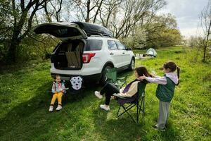 madre con Due figlie sedersi su sedia contro auto Aperto tronco su picnic. foto