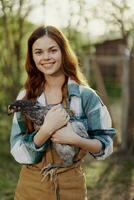 un' contento giovane donna sorridente e Tenere un' giovane pollo quello stabilisce uova per sua azienda agricola foto