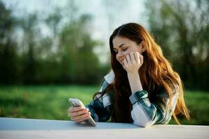 un' giovane femmina blogger si siede all'aperto con sua smartphone nel mano e liberi professionisti nel il all'aperto. il concetto di un' salutare stile di vita e opera foto