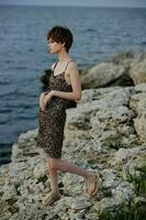 donna nel vestito in piedi su pietre in posa nel spiaggia vestito lusso inalterato foto