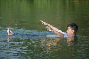 asiatico ragazzo nel bianca maglietta è la spesa il suo tempi liberi di immersione, nuoto, lancio rocce e attraente pesce nel il fiume felicemente, passatempo e felicità di bambini concetto, nel movimento. foto