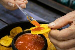 avvicinamento di donna di mani mentre mangiare piantaggine patatine fritte servito con colombiano tradizionale hogao foto