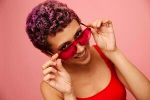 moda ritratto di un' donna con un' corto taglio di capelli nel colorato occhiali da sole e un' Sorridi con denti nel un' rosso superiore su un' rosa sfondo foto